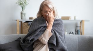 Lady in her 50s, blowing her nose while sitting on the couch with a blanket around her shoulders